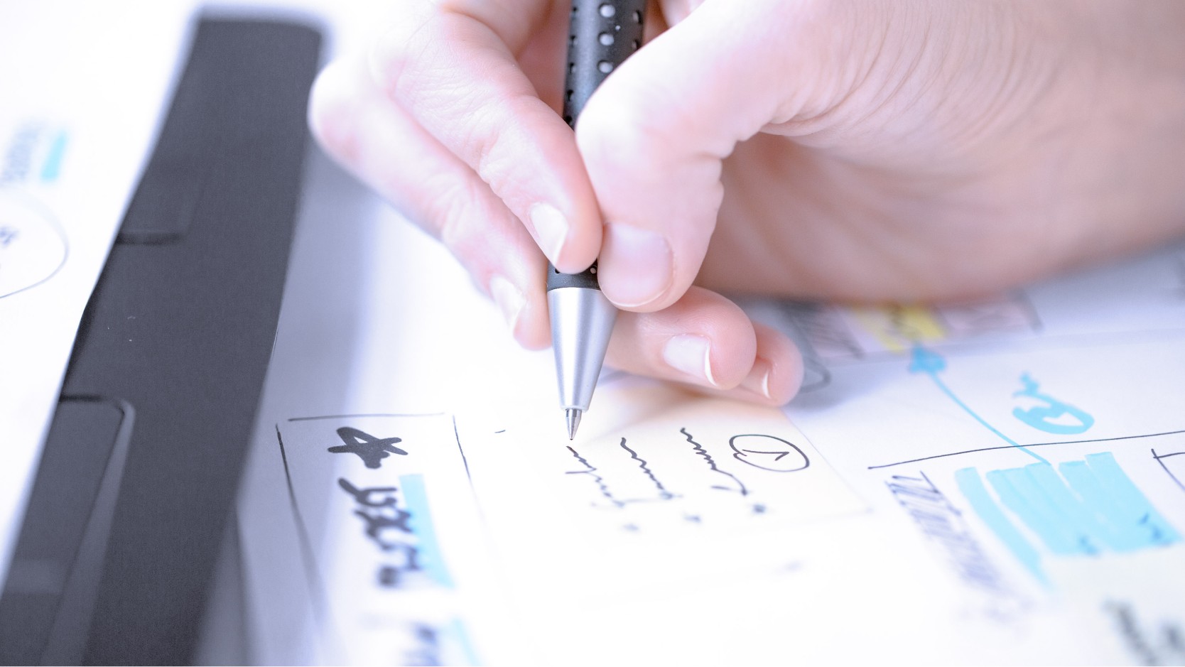 Designer hand holding pen sketching on papers with charts and designs on desk