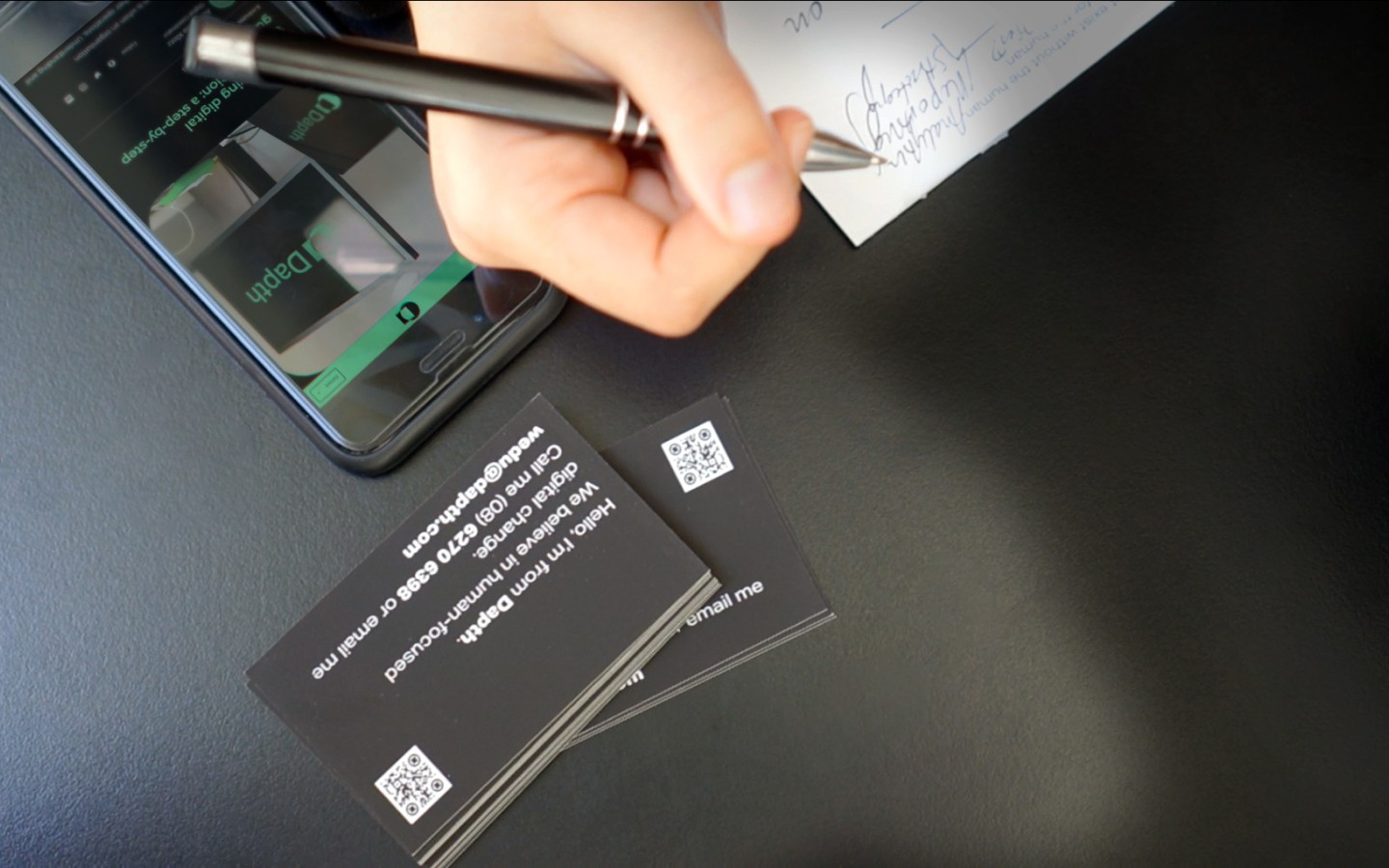 Individual writing on notepad with phone and business cards on table